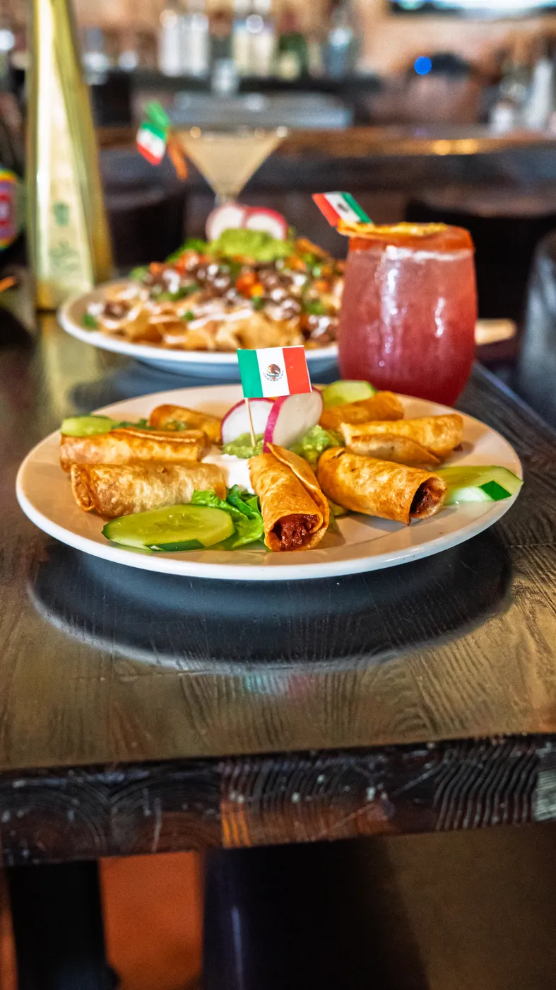 Golden flautas topped with cheese, served with a Mexican flag pick and fresh salsas